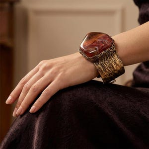 May include: A wide cuff bracelet featuring a large, polished, multi-toned brown and red agate stone set in a textured, dark metallic frame. The band has a hammered, distressed finish with a deep bronze hue, wrapping around the wrist securely.
