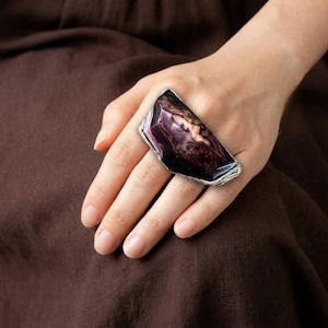 May include: A statement ring featuring a large, rectangular stone with a marbled pattern in shades of purple, brown, and cream. The stone is set in a silver-coloured metal. The ring is displayed on a hand, with a brown fabric background.