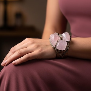 May include: A wide cuff bracelet featuring three large, polished, light pink rose quartz stones set in a textured, dark metallic frame. The stones are arranged in a cluster, showcasing natural variations in the translucent pink mineral surface.