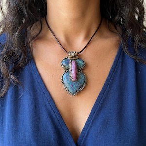 Peut inclure: Un collier pendentif unique avec une grande pierre bleu-vert de forme irrégulière et un accent de pierre rose. Le pendentif est encadré dans un métal doré et est suspendu à un cordon en cuir marron.