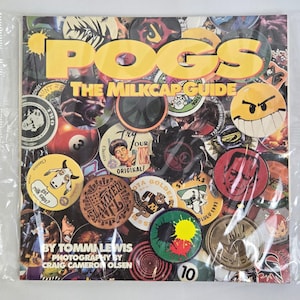 Può includere: Un libro quadrato intitolato "POGS The Milkcap Guide" con una collezione di tappi di latte colorati. La copertina presenta la parola "POGS" in grandi lettere gialle. I tappi di latte hanno vari disegni e testo.