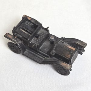 May include: A vintage black toy car with a detailed design, featuring visible seats, wheels, and a front engine compartment. The car has a classic open-top design, with bronze-colored accents on the edges. The toy car is a miniature replica of an antique vehicle.