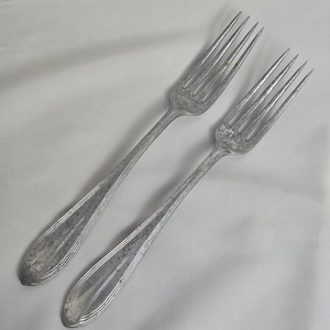 May include: Two vintage silver forks with tines and elongated handles. The forks are made of a silver-coloured metal and have a slightly aged appearance. The handles have a decorative, ridged design. The forks are likely for a formal dining setting.