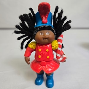 May include: A vintage Cabbage Patch Kids doll dressed as a toy soldier. The doll has dark skin, black yarn hair, and is wearing a red uniform with gold accents, a blue hat, and blue boots. It holds a candy cane.