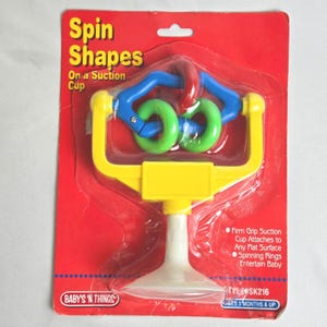 Könnte beinhalten: Ein neues, ungeöffnetes Babyspielzeug namens "Spin Shapes" auf einer roten Kartonrückseite. Das Spielzeug hat einen gelben Rahmen, blaue und grüne Drehringe und einen weißen Saugnapf. Die Verpackung enthält den Text "BABY'S 'N THINGS".