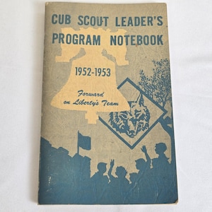 Könnte beinhalten: Vintage Cub Scout Leader's Program Notebook von 1952-1953. Das Cover zeigt eine Glockenillustration mit dem Text "Forward on Liberty's Team". Das Notizbuch hat ein blaues und beiges Farbschema mit Silhouetten und einem Wolfsemblem.