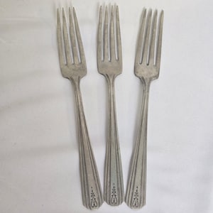May include: Three silver forks with four tines each, arranged on a white surface. The handles have a decorative pattern at the base and vertical lines. The forks are made of a shiny, metallic material.