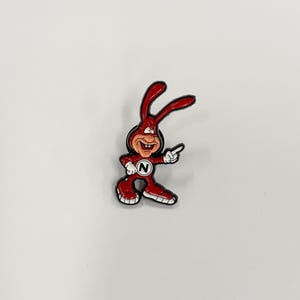 May include: A red cartoon rabbit pin with a white outline. The rabbit is smiling and pointing with its right hand. The rabbit has a white circle with the letter "N" on its chest.