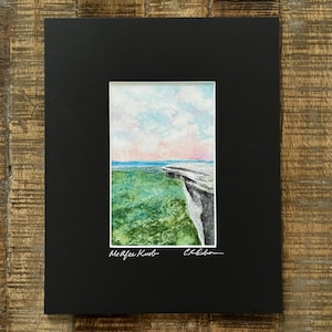 May include: A watercolor painting of a cliff overlooking a green field and a pink and blue sky. The painting is signed "McAfee Kurb" and "C. Gibson".