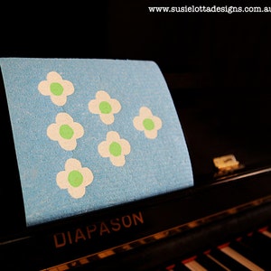 Peut inclure: Un tissu bleu avec des fleurs blanches avec des centres verts. Le tissu est posé sur un piano noir avec le mot "DIAPASON" écrit dessus.