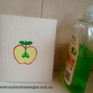 Peut inclure: Une éponge de cuisine blanche avec un motif de pomme rouge. La pomme est coupée en deux et a un centre jaune avec des feuilles vertes. Une bouteille verte de liquide vaisselle avec une étiquette blanche qui dit "Earth Choice" est en arrière-plan.