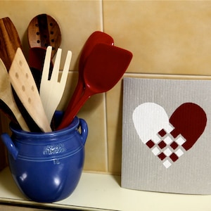 Peut inclure: Un pot en céramique bleu contient une collection d'ustensiles de cuisine en bois, dont une spatule, une fourchette et une cuillère. Une spatule rouge est appuyée contre le pot. Un torchon gris avec un motif de cœur blanc et rouge est sur le comptoir.