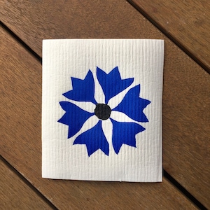 Peut inclure: Un carré de tissu blanc avec un motif floral bleu audacieux. La fleur a des pétales pointus et un centre noir. Le tissu est posé sur une surface en bois.