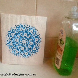 Peut inclure: Une éponge de cuisine blanche avec un motif de dentelle bleue imprimé dessus. L'éponge est posée sur un comptoir en carrelage blanc à côté d'une bouteille en plastique transparente de liquide vaisselle vert.