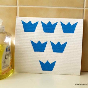 Peut inclure: Un torchon blanc avec des couronnes bleues imprimées dessus. Le torchon est accroché à un mur carrelé à côté d'une bouteille transparente de liquide jaune.