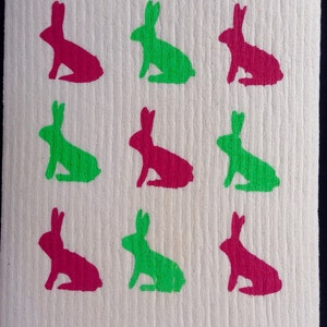 Peut inclure: Une éponge de cuisine blanche avec neuf silhouettes de lapins imprimées dessus. Les lapins alternent entre le vert et le rose.