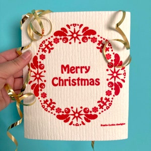 Peut inclure: Un torchon blanc avec un motif de couronne rouge qui dit "Joyeux Noël" au centre. La couronne est composée d'étoiles stylisées, de baies et de cœurs. Le torchon est décoré d'un ruban doré.