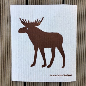 Peut inclure: Une éponge à vaisselle en coton blanc avec une silhouette de moose marron. L'éponge a une surface texturée et le moose a un œil blanc. Le texte "Susie-Lotta Designs" est imprimé en marron au bas de l'éponge.