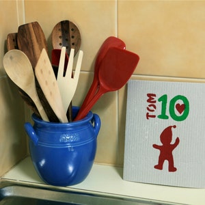Peut inclure: Un pot en céramique bleu contient une collection d'ustensiles de cuisine en bois, dont une cuillère en bois, une spatule en bois et une fourchette en bois. Une spatule en plastique rouge est appuyée contre le pot. Un torchon blanc avec le texte "TOM 10" et un cœur rouge est sur le comptoir.
