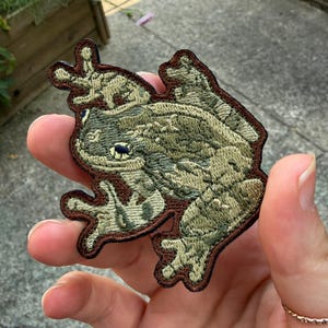 May include: An embroidered patch shaped like a frog with textured light green and olive green thread detailing. The frog has a dark brown stitched border outlining its body and limbs, with a smaller frog figure positioned on its back.