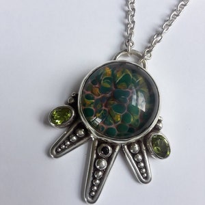 May include: A silver pendant necklace with a green and gold glass cabochon set in a silver starburst design. The pendant is accented with four green gemstones.