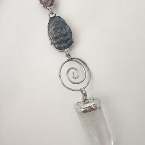 May include: A handcrafted pendant featuring a clear quartz crystal point, a carved blue-grey Buddha head, and a purple stone. The pendant is connected by silver-toned metal and a spiral design. The pendant hangs from a thin, clear cord.