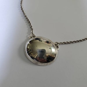 Op de afbeelding: Een zilveren ketting met een grote, ronde hanger. De hanger is glad en reflecterend en hangt aan een delicate ketting.