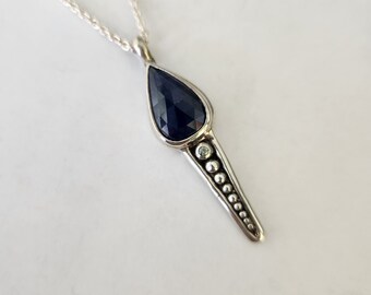 May include: A silver necklace with a teardrop-shaped pendant featuring a dark blue gemstone and a row of silver beads.