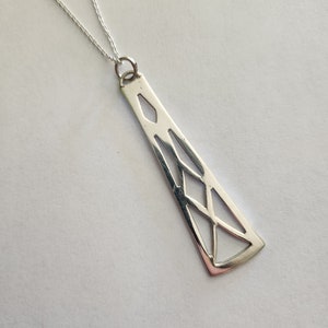 Op de afbeelding: Een zilveren ketting met een geometrische hanger. De hanger is een lange, dunne rechthoek met een uitgesneden ontwerp.