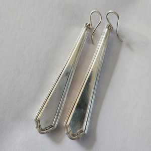 May include: A pair of silver-colored, elongated earrings with a rectangular shape and a decorative edge. The earrings have a hook closure and are displayed on a white background. The earrings are a jewelry item.
