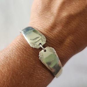 May include: A silver bracelet made from repurposed silverware. The bracelet features two rectangular pieces connected by a small chain. Each piece has a decorative edge and a reflective surface. The bracelet is worn on a wrist.