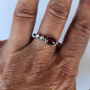 Könnte beinhalten: Ein silberner Ring mit einem roten ovalen Edelstein und zwei kleineren runden weißen Edelsteinen.