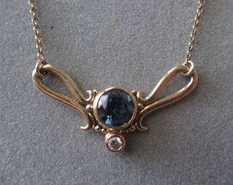 Op de afbeelding: Een gouden ketting met een blauwe edelsteen hanger en een kleinere, heldere edelsteen. De hanger is gezet in een delicaat, versierd ontwerp.
