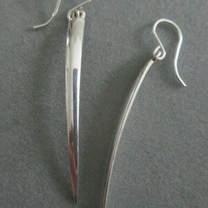 Könnte beinhalten: Ein Paar silberne Ohrringe mit einem langen, dünnen, spitzen Design. Die Ohrringe sind aus glattem, poliertem Metall.