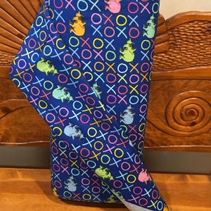 May include: A folded fabric roll featuring a vibrant blue background with a repeating pattern of colorful X's, O's, and cartoon characters. The characters are in shades of yellow, green, pink, and gray. The fabric is displayed on a wooden bench.