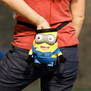May include: A woman wearing a red tank top and blue jeans is holding a yellow and blue Minion-themed chalk bag.