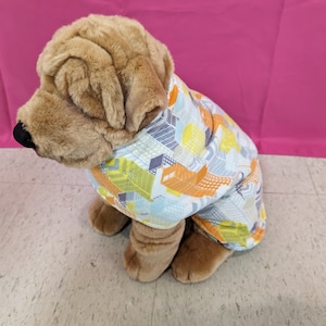 May include: A brown and tan plush dog wearing a blue, yellow, and orange patterned shirt. The dog is sitting on a light gray tiled floor.