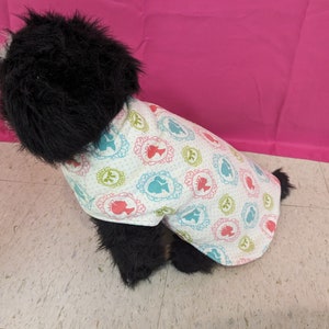 May include: A black and white dog wearing a white dog shirt with a pattern of colorful silhouettes of women in frames.