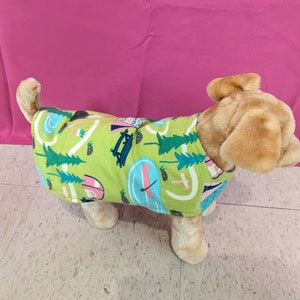May include: A stuffed dog wearing a green and blue patterned dog shirt with a camping theme. The shirt has images of trees, tents, and a lake.