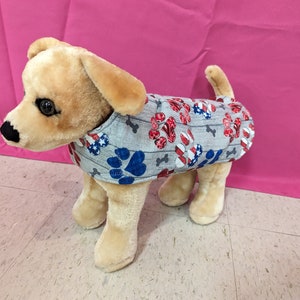 May include: A plush dog wearing a gray dog shirt. The shirt is patterned with red, white, and blue paw prints, stars, and bones. The dog is light brown with black eyes and a pink background.