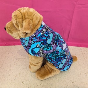 May include: A brown and tan plush dog wearing a blue and purple dog sweater with a colorful print of hearts, stars, and flowers.