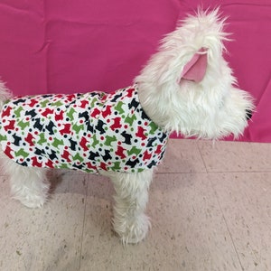 May include: A white dog wearing a red, green, and black patterned dog coat with a white background. The coat has a repeating pattern of small black dogs and red and green polka dots.