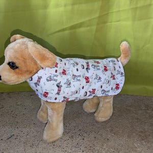 May include: A plush dog toy in tan and cream colors, wearing a white coat patterned with red and gray dog illustrations. The toy has a black nose and dark eyes, and is standing on a gray surface.