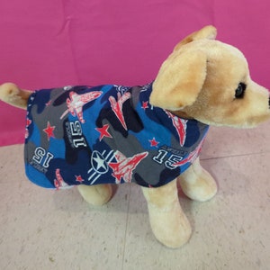 May include: A small, light brown dog wearing a blue and gray dog shirt with a camouflage pattern, red stars, and the text "US Air Force" and "15" on it.