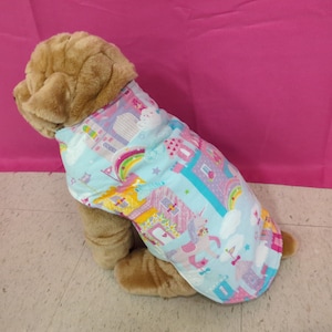 May include: A light blue dog vest with a colorful unicorn, rainbow, and castle print. The vest is worn by a light brown dog.