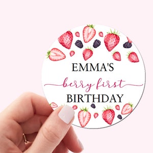 May include: A white round sticker with a red and black berry design. The text "EMMA'S berry first BIRTHDAY" is printed on the sticker.