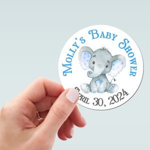 Pu&ograve; includere: Un adesivo rotondo con un'illustrazione di elefante blu e bianco. Il testo "Molly's Baby Shower" &egrave; scritto in blu intorno all'elefante. La data "April 30, 2024" &egrave; scritta in nero sotto l'elefante.