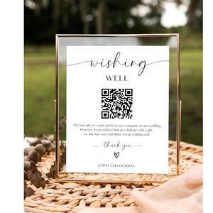 May include: A white sign with black text and a QR code in a gold frame. The sign reads "wishing well" and includes a message asking guests to contribute to a wishing well. The sign is on a woven wicker surface.