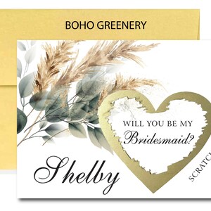 May include: A white card with a gold heart that says "Will you be my Bridesmaid?" with a scratch-off area. The card has a watercolor design of greenery and pampas grass. The name "Shelby" is written in cursive at the bottom of the card.