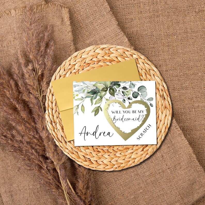 May include: A white card with green watercolor leaves and a gold heart with the text "Will you be my bridesmaid?"  The card has the name "Andrea" printed on it and the word "Scratch" below the heart.